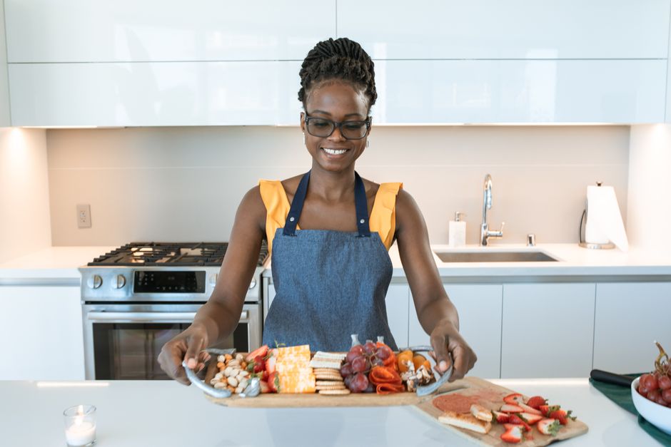 Smiling woman in kitchen with charcuterie board featuring cheese, meats, and fruits.