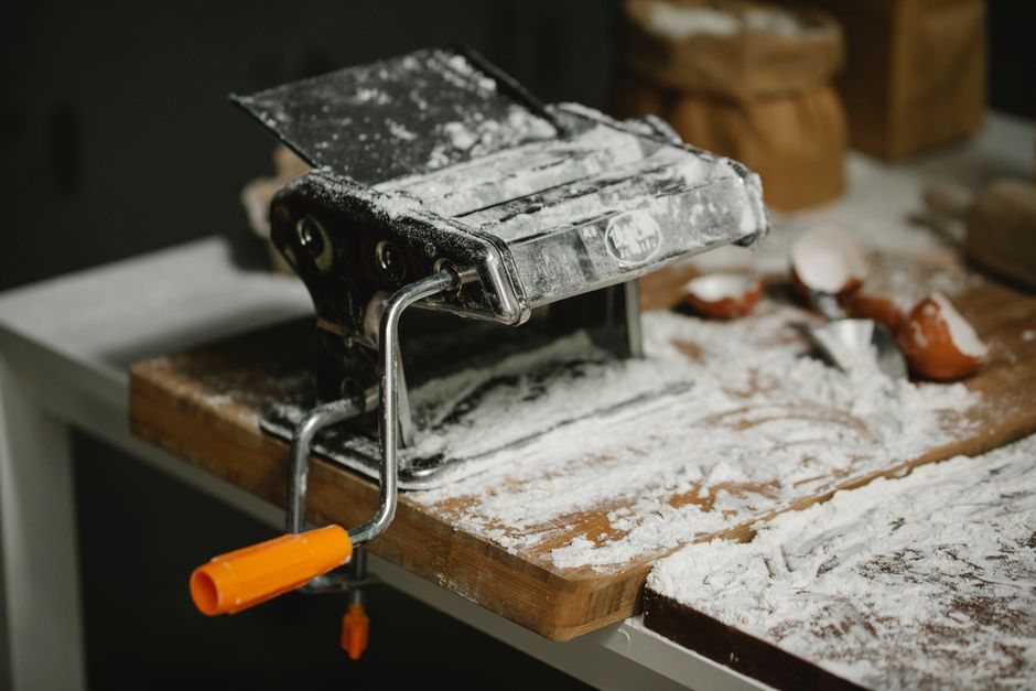 Modern manual pasta roller and cutter machine placed on wooden surface covered with flour in kitchen
